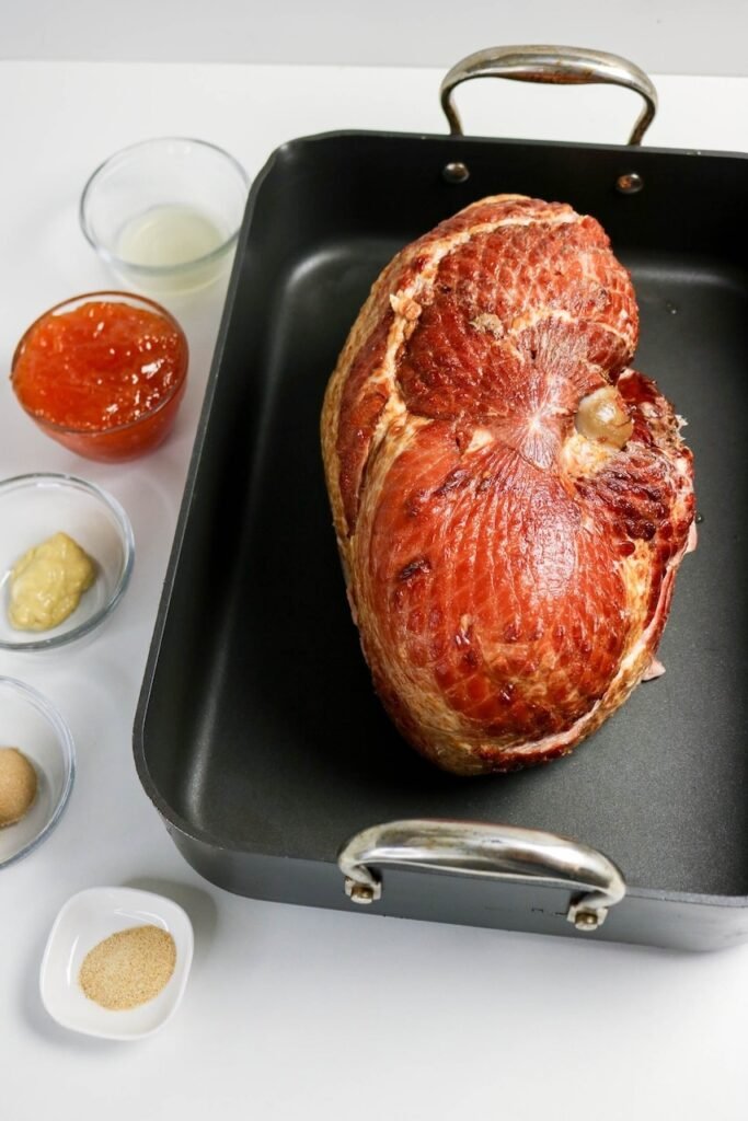 Spiral-cut pork placed in a roasting pan with bowls of apricot preserves, Dijon mustard, brown sugar, apple cider vinegar, and garlic powder ready.
