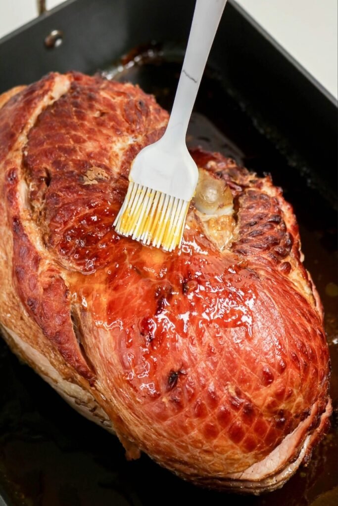 Brushing apricot glaze over a baked ham using a pastry brush to create a glossy, caramelized crust during the recipe.