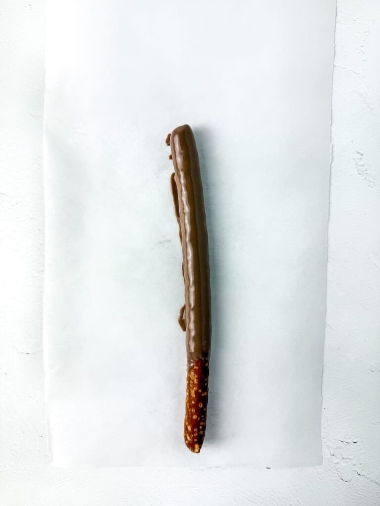 Step four showing a single pretzel rod freshly dipped in melted chocolate and placed on a sheet of parchment paper to set. The chocolate coating is smooth and even, forming the base layer for decorating.