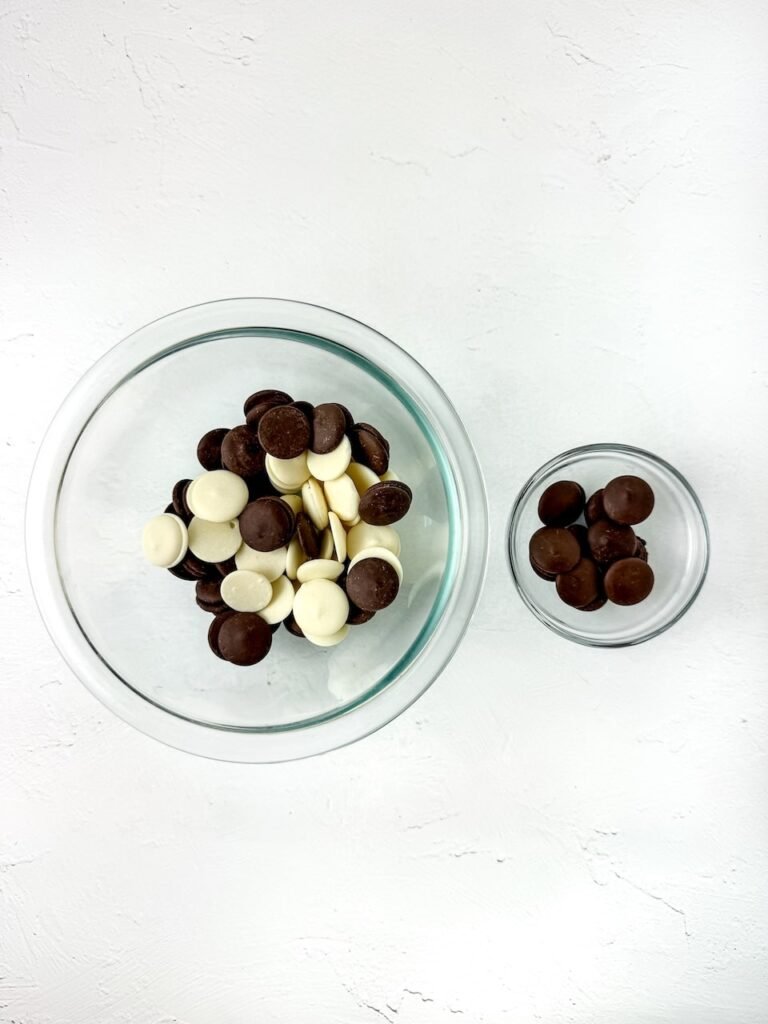 Step one in the process showing two glass bowls filled with chocolate and white candy melts before being melted. The ingredients are arranged on a white surface, ready for dipping pretzel rods in the next step.
