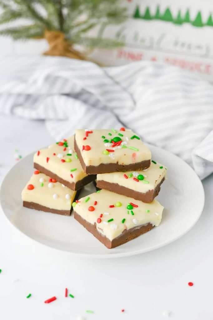 Stacked pieces of layered Christmas chocolate fudge with white chocolate tops, festive red and green sprinkles, and rich chocolate bottoms on a white plate.