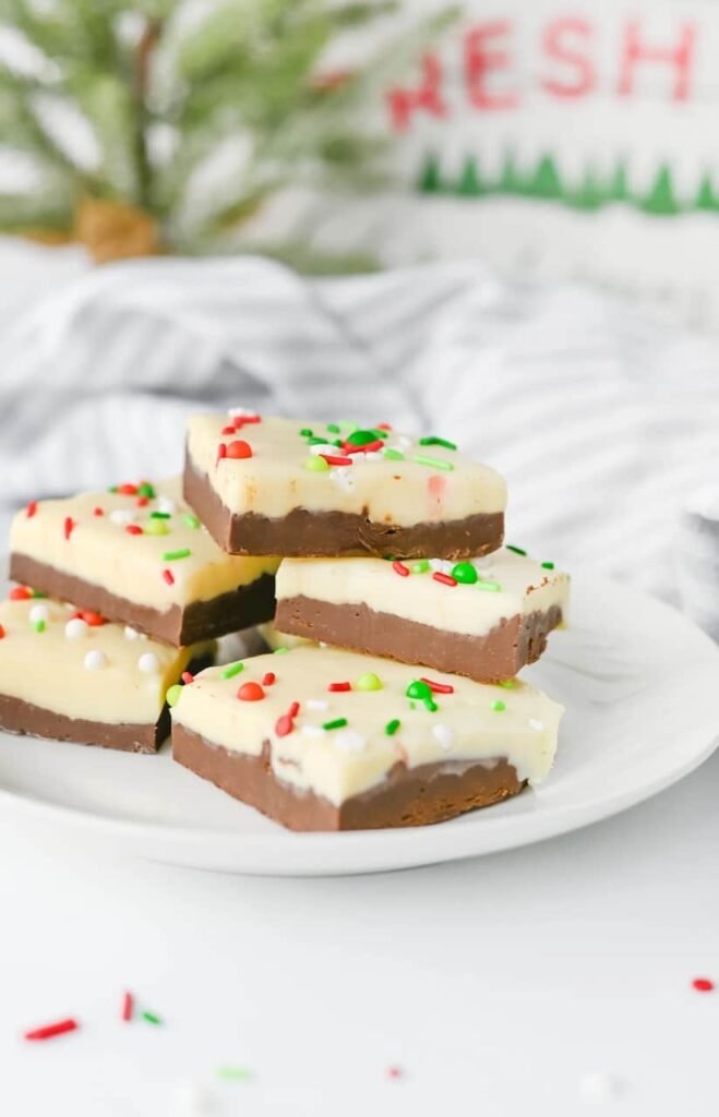 Stacked pieces of layered Christmas chocolate fudge with white chocolate tops and festive sprinkles on a white plate in front of a Christmas background.