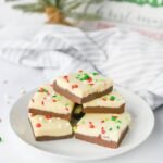 Layered Christmas chocolate fudge with white and milk chocolate layers topped with red and green sprinkles on a plate in front of a small Christmas tree.