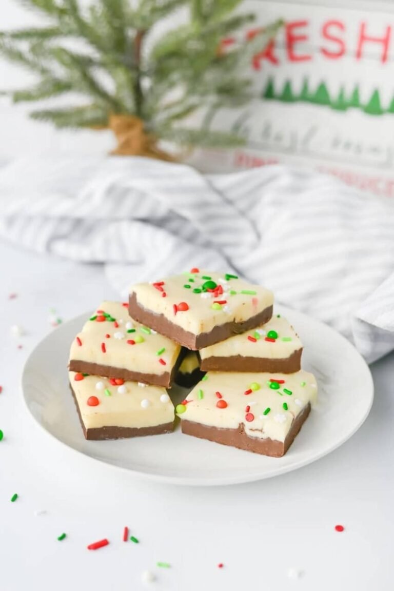 Layered Christmas chocolate fudge with white and milk chocolate layers topped with red and green sprinkles on a plate in front of a small Christmas tree.
