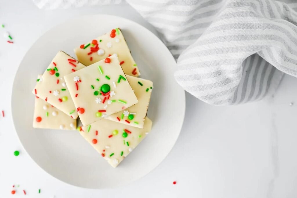 Top view of layered Christmas chocolate fudge squares with white chocolate tops and festive red and green sprinkles on a white plate.