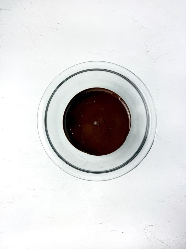 A glass bowl filled with melted brown chocolate on a white surface, prepared for coating Nutter Butter cookies while making no-bake owl cookies for a fall dessert recipe.