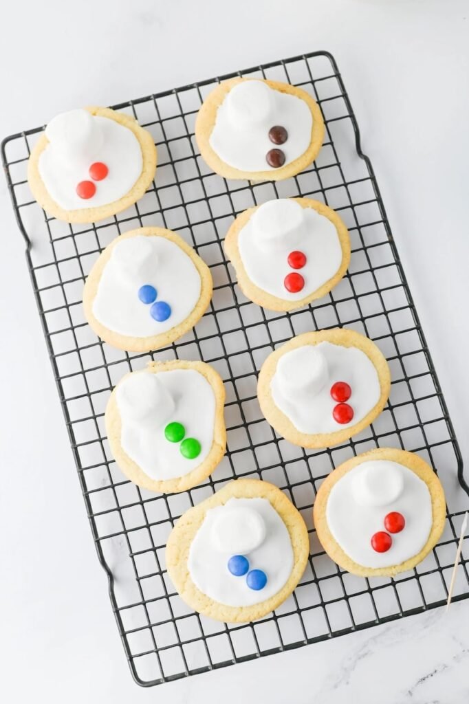 Partially decorated melted snowman cookies with marshmallow heads and colorful candy buttons cooling on a wire rack.