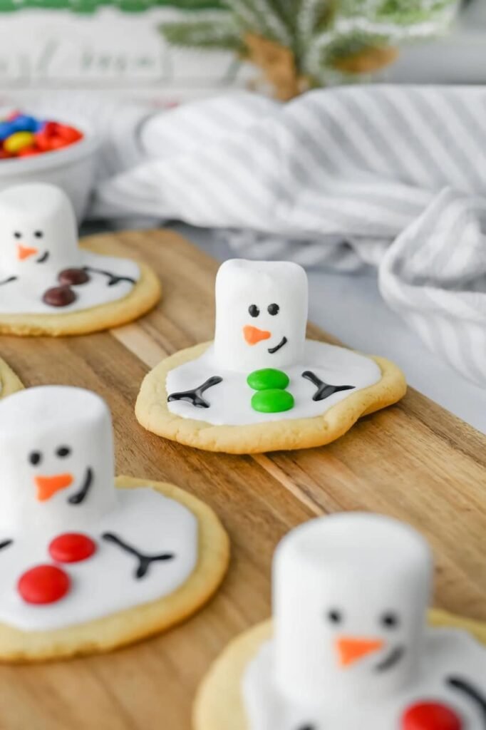 Melted snowman cookies decorated with marshmallow heads, icing arms, and colorful candy buttons on a wooden board with holiday decor in the background.