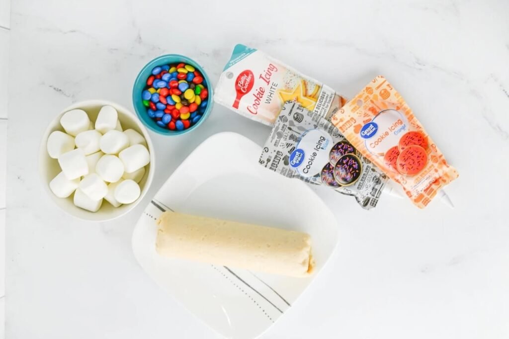 Ingredients for melted snowman cookies including marshmallows, M&M candies, sugar cookie dough, and cookie icing tubes on a white background.