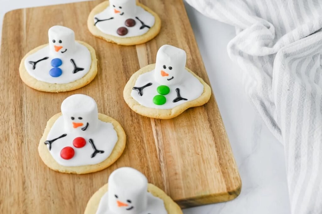 Melted snowman cookies decorated with marshmallow heads, icing, and candy buttons arranged on a wooden board.