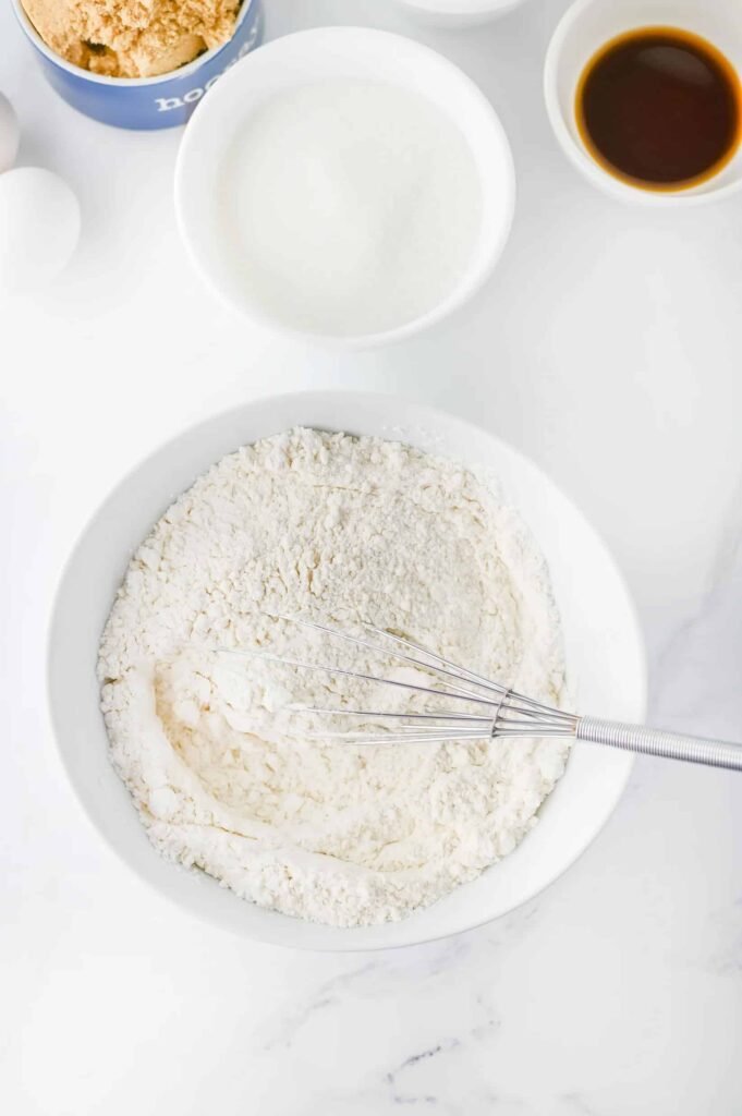 Whisk mixing dry ingredients for peppermint chocolate chip cookies in a white bowl on a marble countertop.