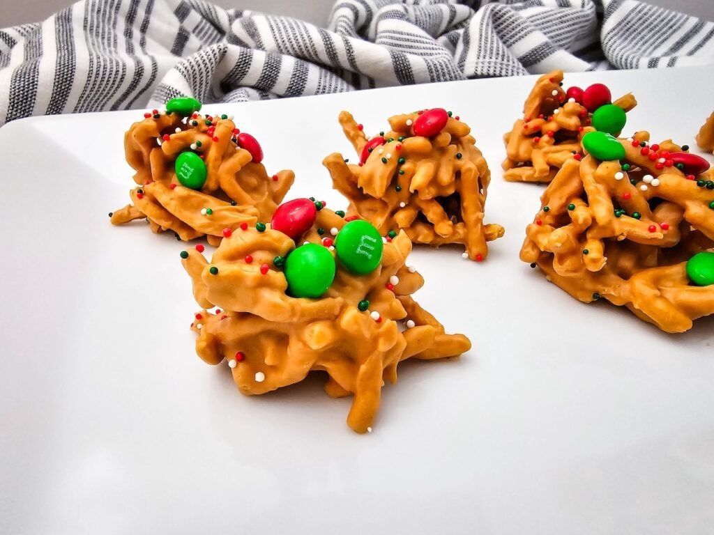 No-bake Christmas haystack cookies made with butterscotch, chow mein noodles, and decorated with red and green M&Ms and holiday sprinkles on a white plate.
