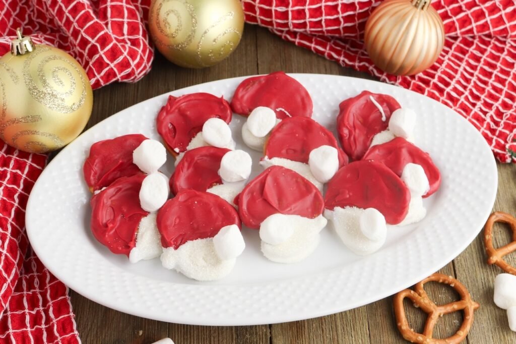 A white platter of red and white Pretzel Santa Hats topped with mini marshmallows, surrounded by gold ornaments and a festive red towel.