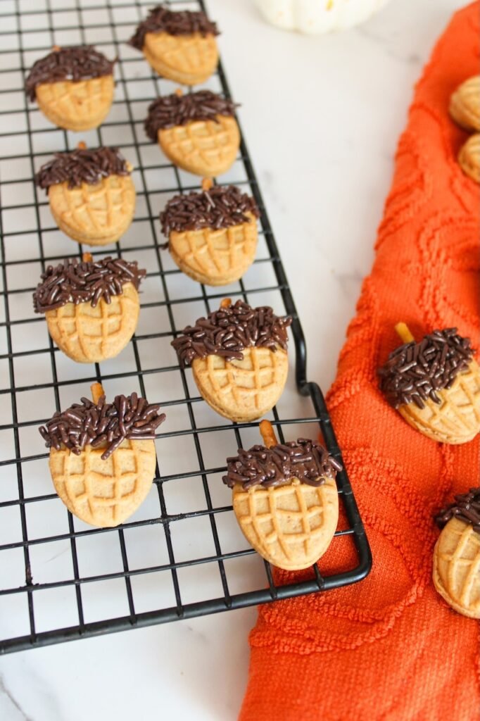 Close-up of Nutter Butter acorn cookies topped with chocolate sprinkles and pretzel stick stems cooling on a wire rack beside an orange fall towel, ready to serve as an easy no-bake fall dessert.