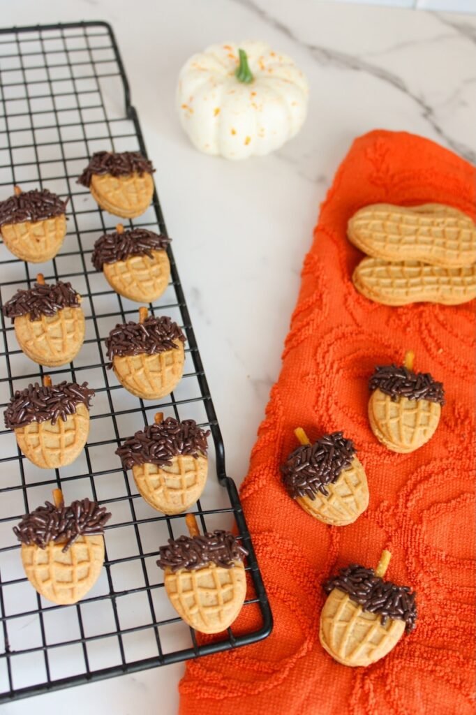 Overhead view of Nutter Butter acorn cookies with chocolate sprinkle tops and pretzel stick stems cooling on a wire rack beside an orange towel and a small white pumpkin, styled as a cozy fall dessert scene.