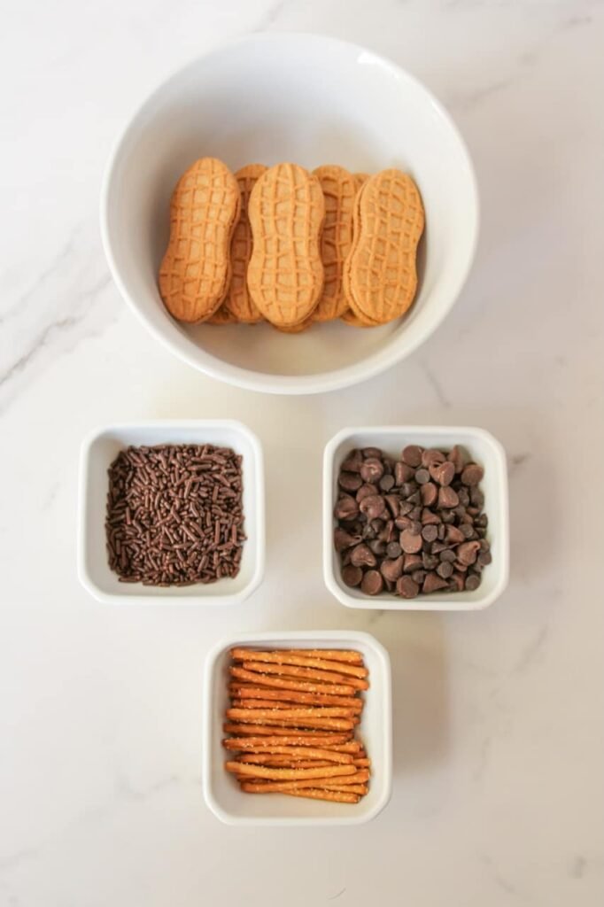 Overhead view of Nutter Butter acorn ingredients arranged on a white marble surface, including a bowl of peanut-shaped Nutter Butter cookies, small dishes of chocolate sprinkles, milk chocolate chips, and pretzel sticks used for the acorn stems.