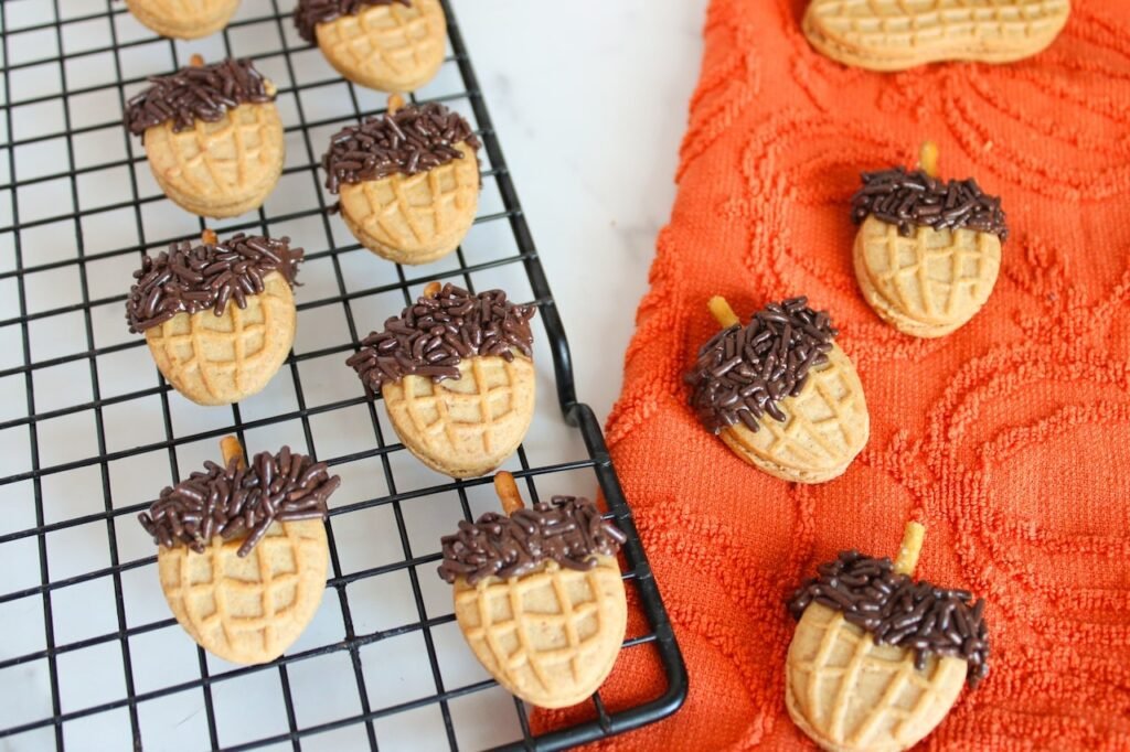 Freshly made Nutter Butter acorn cookies with chocolate sprinkle tops and pretzel stick stems cooling on a wire rack beside an orange fall towel, ready to serve as an easy no-bake fall treat.