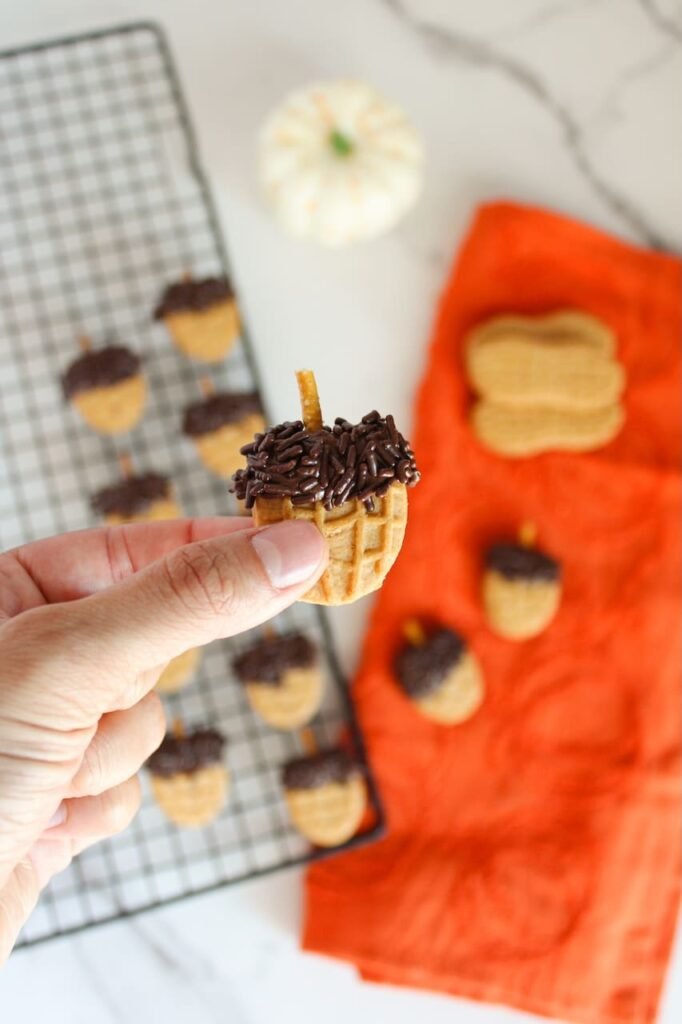 Close-up of a hand holding a finished Nutter Butter acorn cookie with chocolate sprinkles and a pretzel stick stem, with more acorns on a cooling rack and orange towel in the background for a cozy fall presentation.