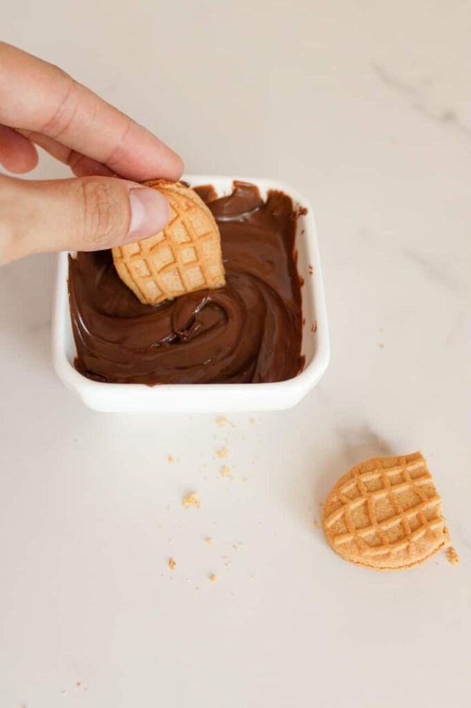 Hand dipping half into a small dish of melted chocolate, demonstrating step three of making Nutter Butter acorns on a white countertop.