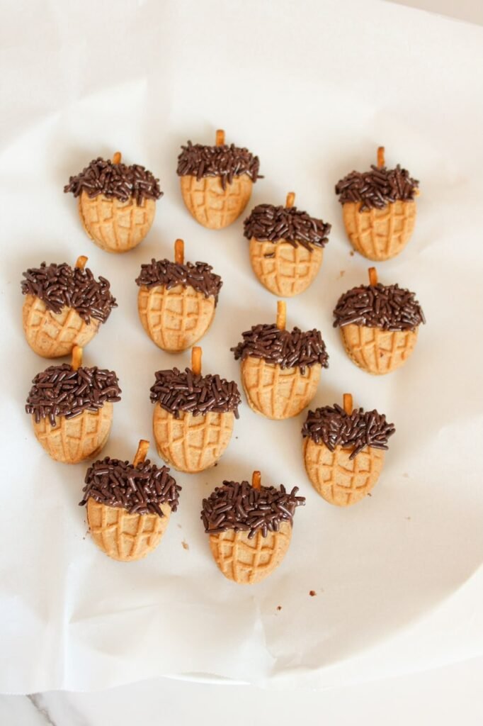 Overhead view of multiple finished Nutter Butter acorn cookies on parchment paper, each topped with chocolate sprinkles and a pretzel stick stem, cooling and setting after assembly.