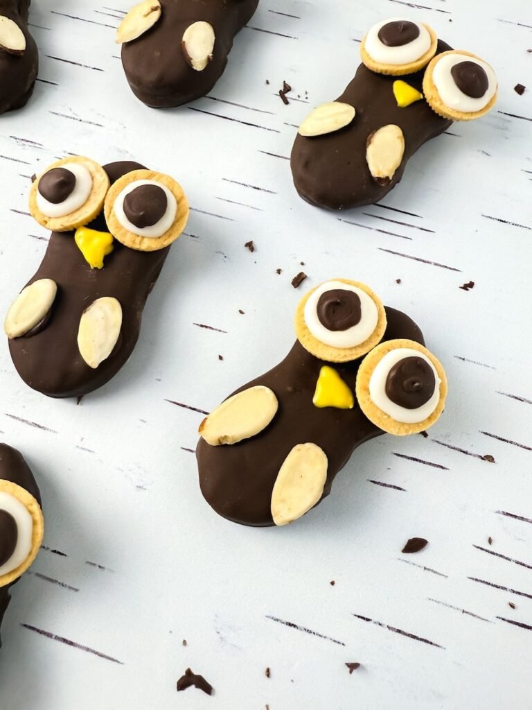 Angled view of Nutter Butter owl cookies on a white birch-patterned background, each coated in melted chocolate and decorated with cookie eyes, yellow beaks, and almond slice wings, shown as an easy no-bake fall dessert.