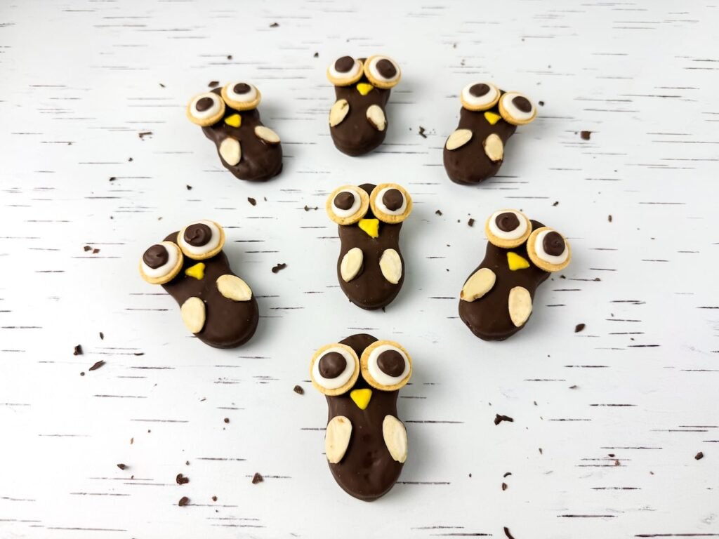 Several Nutter Butter owl cookies arranged on a white birch-patterned surface, each coated in melted chocolate and decorated with round cookie eyes, yellow candy beaks, and almond slice wings.