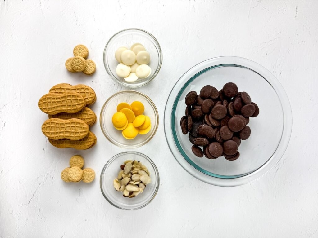Ingredients for Nutter Butter owl cookies including peanut-shaped cookies, candy melts, mini sandwich cookies, and slivered almonds arranged in bowls on a white background.