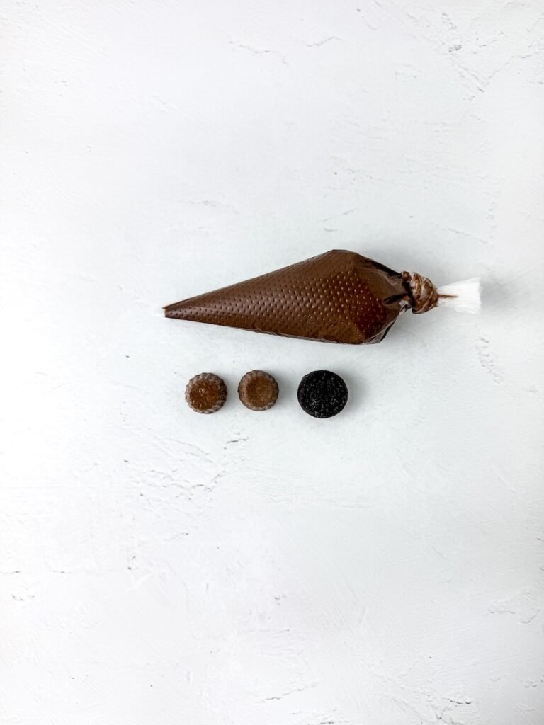 Step two in the process showing a piping bag filled with melted chocolate next to two mini peanut butter cups and a mini Oreo cookie on a white background. This setup is used to assemble the scarecrow hats before attaching them to the dipped pretzels.