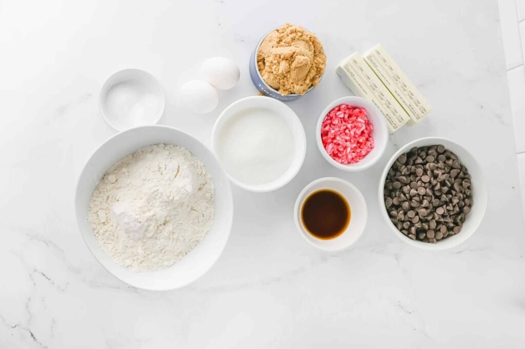 Ingredients for peppermint chocolate chip cookies including butter, sugar, flour, eggs, vanilla, chocolate chips, and crushed candy canes arranged on a white counter.