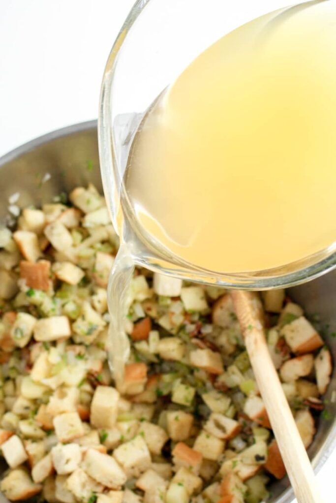Pouring broth into a bowl of bread cubes, apples, celery, and herbs to make moist and flavorful herb and apple stuffing.