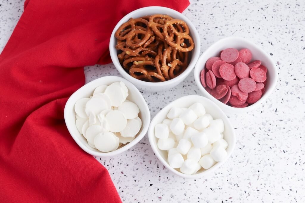 Bowls of mini pretzels, red and white candy melts, and mini marshmallows on a white countertop with a red cloth, ingredients for Pretzel Santa Hats.