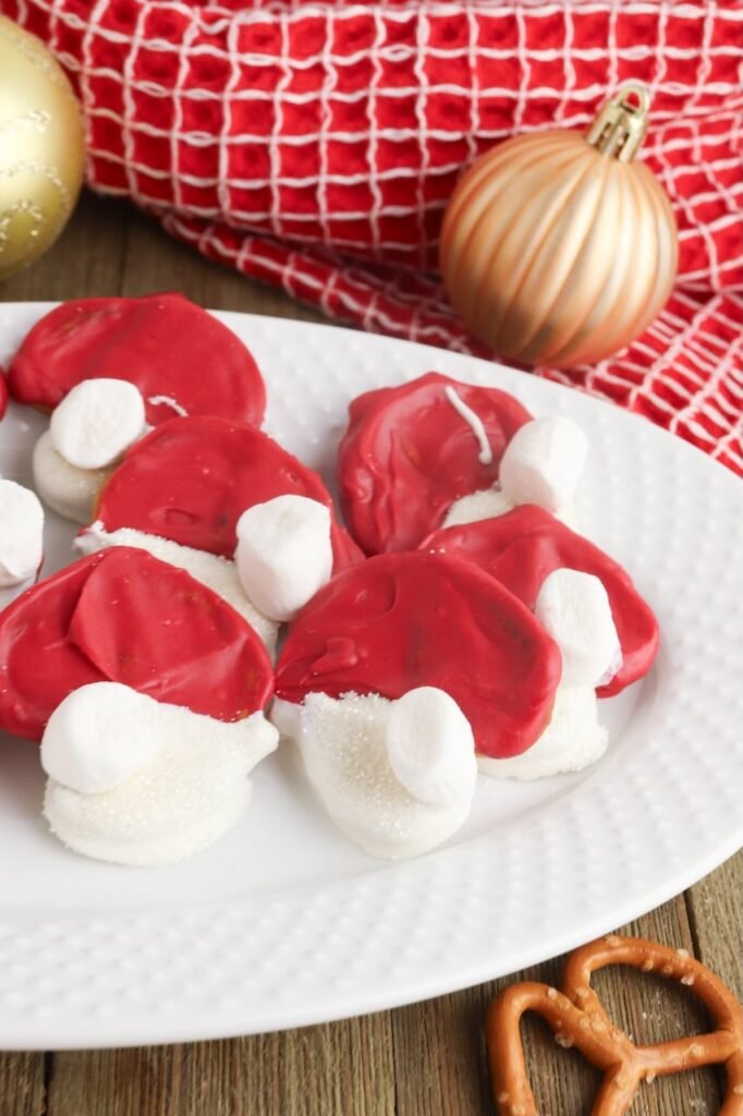 Vertical photo of Pretzel Santa Hats made with red and white chocolate and mini marshmallows on a white plate, styled with holiday ornaments and a red towel.