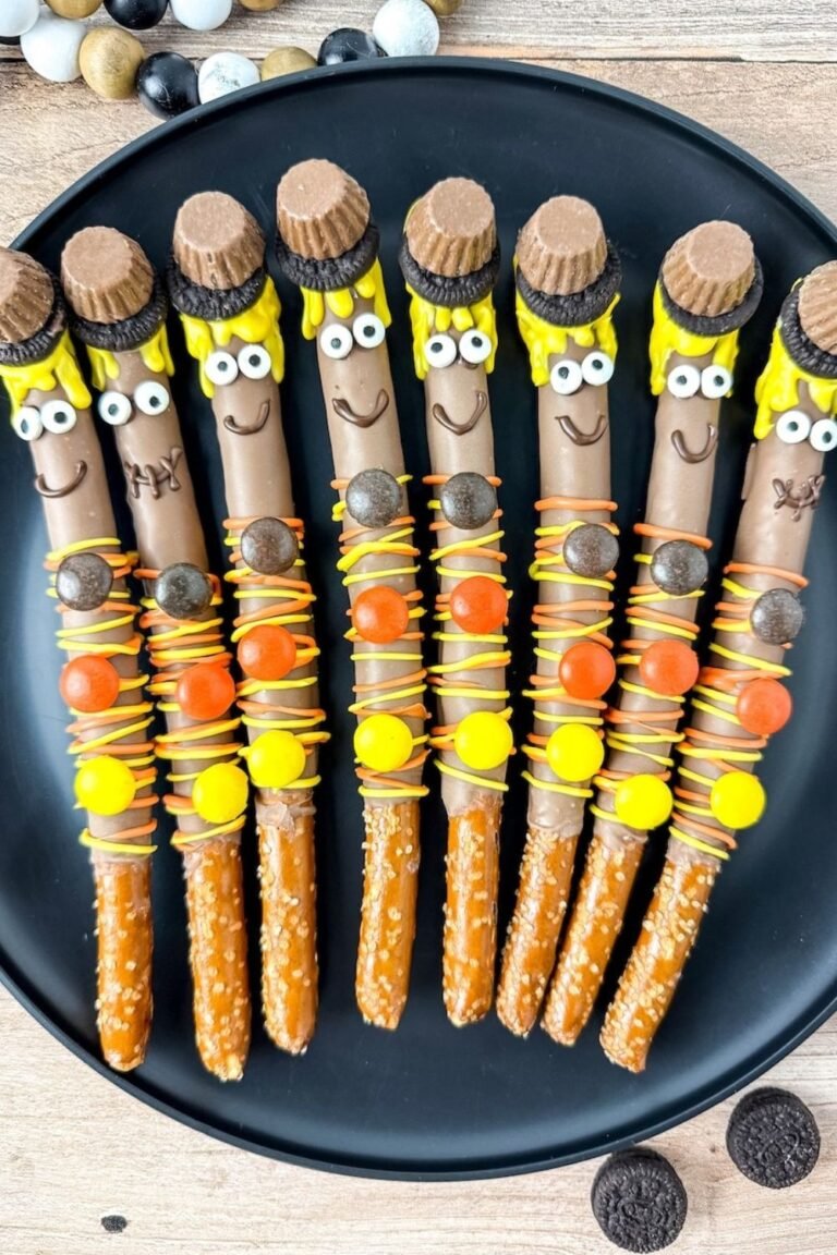 Vertical image showing a close-up of finished scarecrow pretzel rods on a black plate. Each pretzel is dipped in chocolate, decorated with candy eyes, yellow candy melt hair, peanut butter cup and Oreo hats, and topped with orange and yellow candy drizzle and Reese’s Pieces buttons. A perfect no-bake fall dessert or Halloween treat for kids.