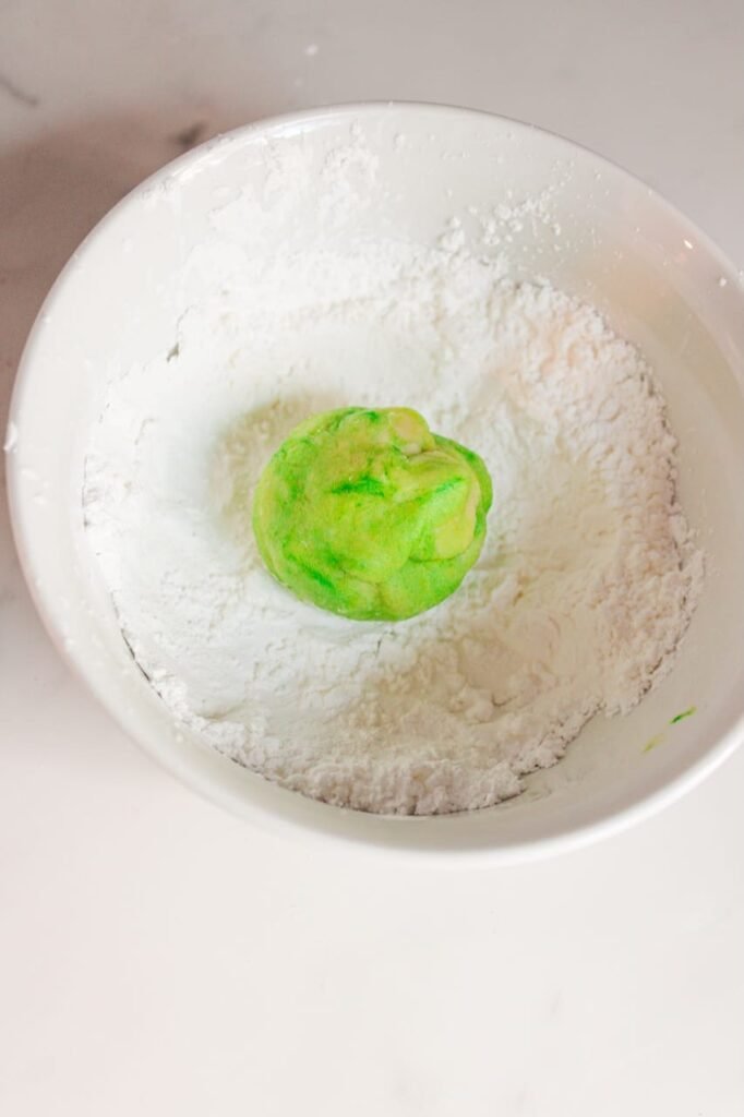 Bright green cookie dough ball being rolled in powdered sugar to create a snowy coating for holiday cookies.