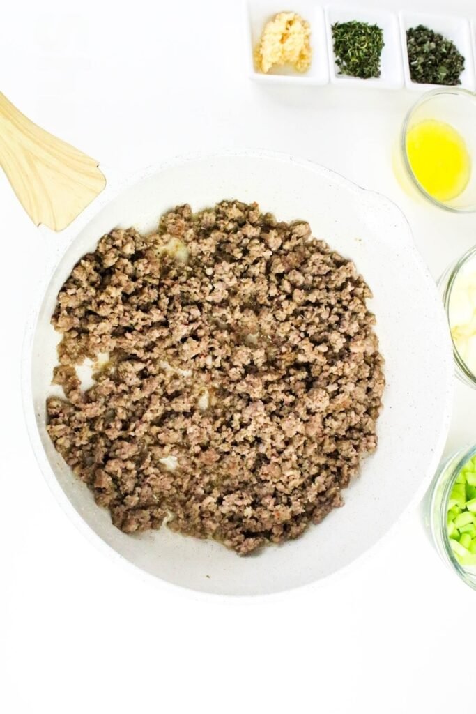 Browned ground sausage cooking in a white skillet with garlic, herbs, and melted butter nearby.