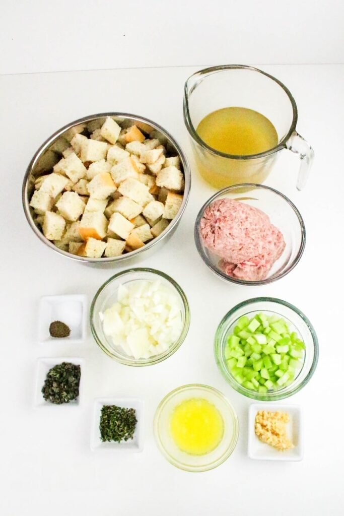 Ingredients for sage and sausage herb stuffing including cubed bread, ground sausage, celery, onion, broth, melted butter, garlic, and fresh herbs on a white background.