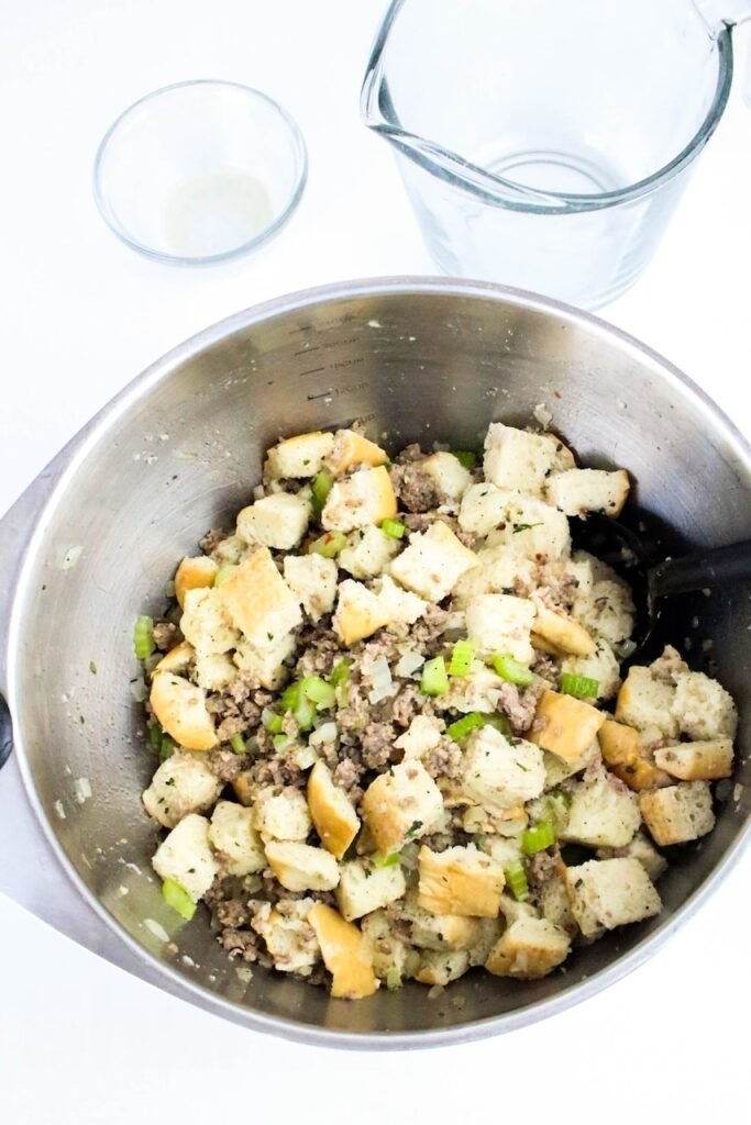 Mixing cubed bread, cooked sausage, celery, and onions in a large stainless steel bowl to make sage and sausage herb stuffing.