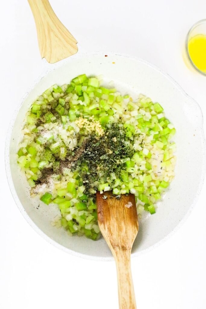 Chopped celery, onion, garlic, and fresh herbs sautéing in a white skillet with a wooden spoon.
