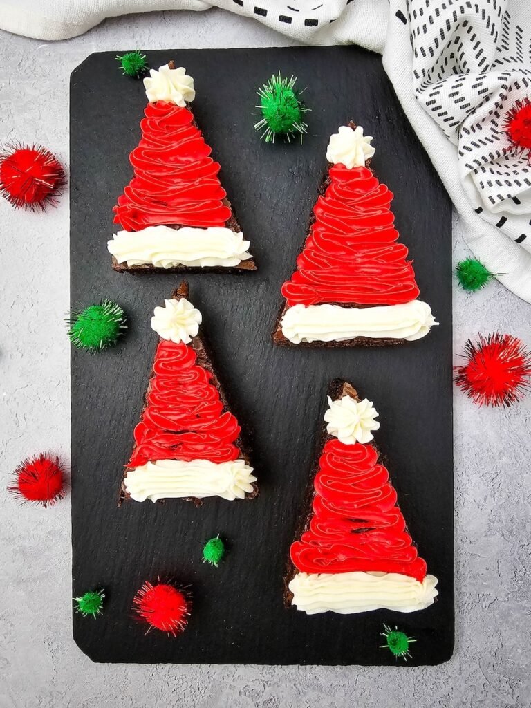 Four decorated Santa Hat Brownies with red and white frosting arranged on a slate board with holiday pom-poms.