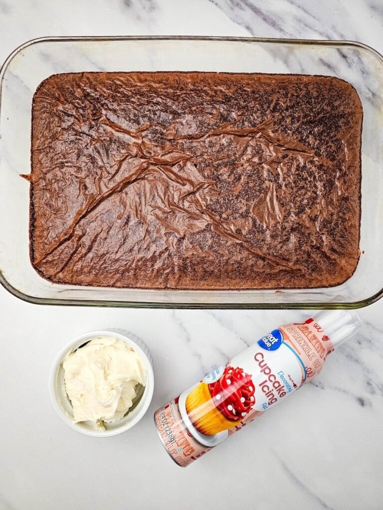 Pan of baked brownies, vanilla frosting, and red cupcake icing on marble surface.