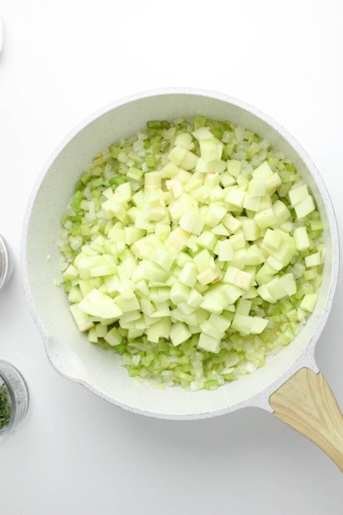 Diced apples, celery, and onions sautéing in butter in a white skillet.