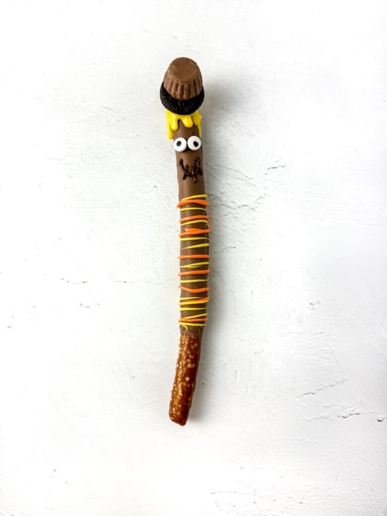 Close-up of a finished scarecrow pretzel rod decorated with a mini peanut butter cup and Oreo hat, candy eyes, yellow candy melt hair, and orange and yellow drizzle for the scarf. The pretzel is set on a white background, showing a cute and creative no-bake fall dessert idea.