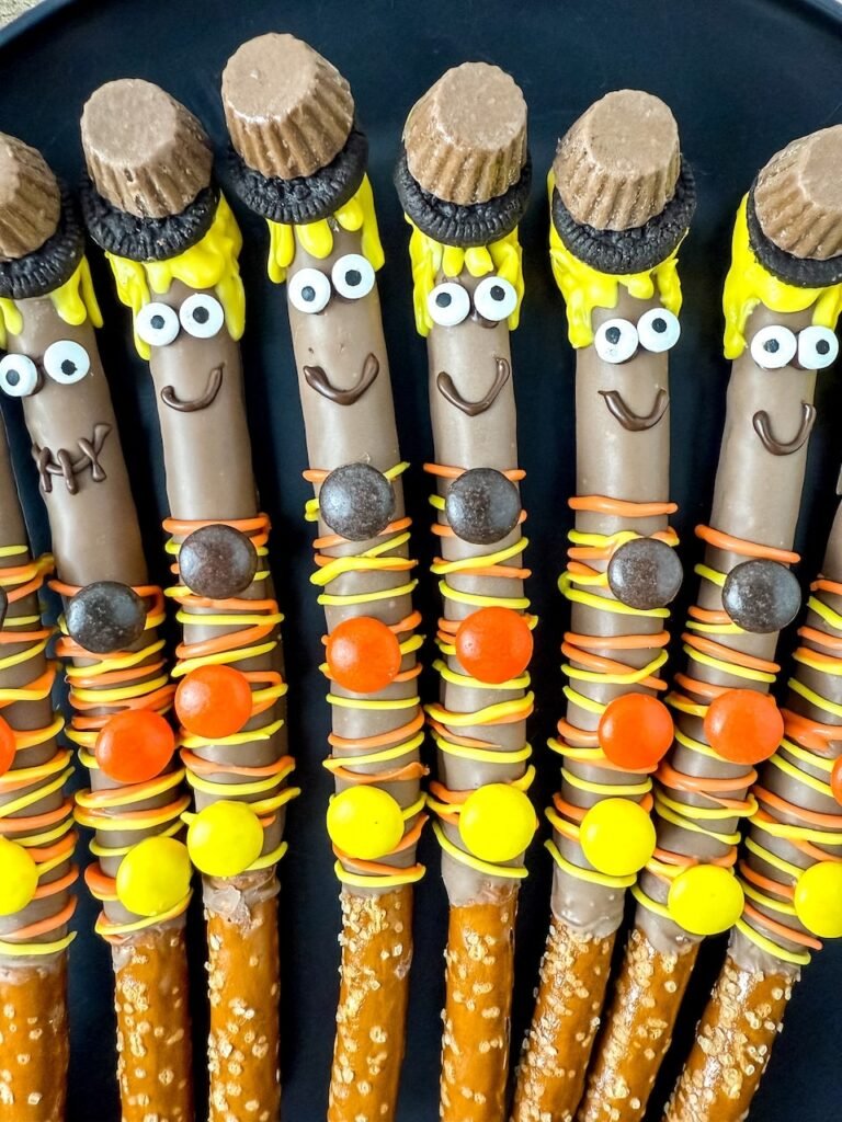 Close-up of several finished scarecrow pretzel rods arranged side by side on a dark plate. Each pretzel is dipped in chocolate and decorated with yellow candy melt hair, candy eyes, peanut butter cup and Oreo hats, and orange and yellow drizzle with Reese’s Pieces buttons. A festive no-bake fall dessert perfect for kids and parties.