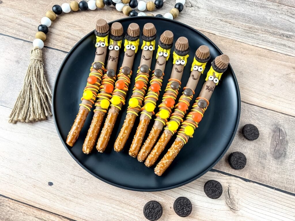 A group of finished scarecrow pretzel rods arranged neatly on a black plate over a rustic wooden background. Each pretzel is coated in chocolate and decorated with yellow candy melt hair, candy eyes, Oreo and peanut butter cup hats, and colorful Reese’s Pieces buttons. The festive fall dessert is surrounded by mini Oreos and a farmhouse-style beaded garland for cozy seasonal presentation.