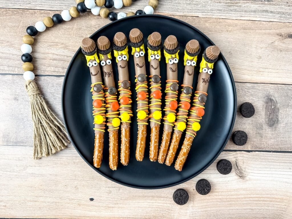 A set of homemade scarecrow pretzel rods arranged on a black plate, decorated with yellow candy melt “hair,” mini peanut butter cup hats, candy eyes, and orange and yellow Reese’s Pieces buttons. The chocolate-dipped pretzels are displayed on a wooden surface with mini Oreo cookies and a rustic fall garland for decoration.