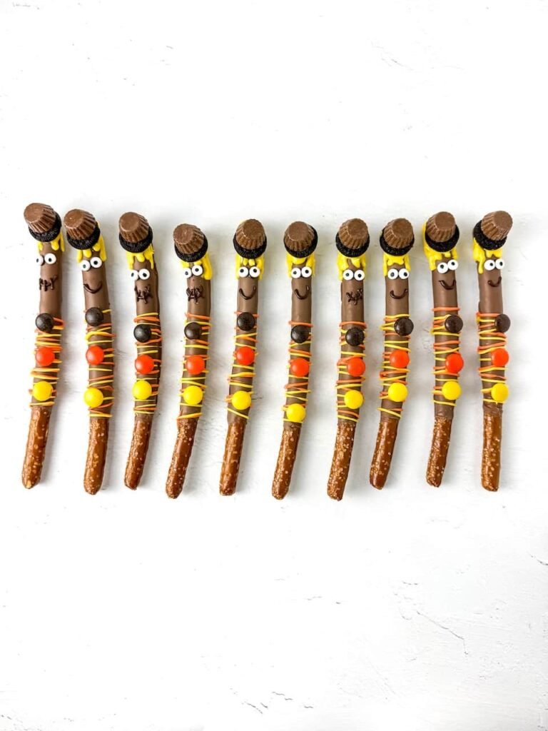 A row of finished scarecrow pretzel rods decorated with chocolate coating, yellow candy melt “straw” hair, candy eyes, peanut butter cup and Oreo hats, and Reese’s Pieces buttons. The colorful fall treats are arranged neatly on a white background, ready for serving or gifting.