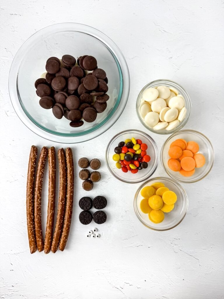 Overhead view of ingredients to make scarecrow pretzel rods, including pretzel sticks, chocolate melting wafers, peanut butter cups, mini Oreos, Reese’s Pieces, candy eyes, and colored candy melts in orange, yellow, and white bowls arranged on a white background.