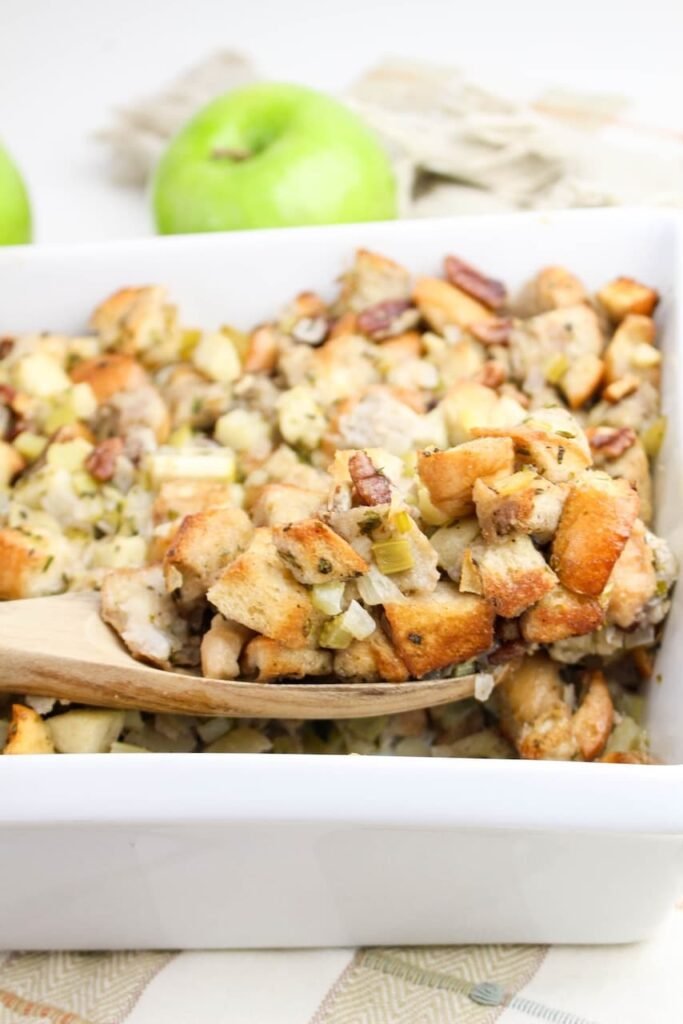 A wooden spoon scooping baked herb and apple stuffing from a white casserole dish, showing golden bread cubes, apples, celery, and pecans.