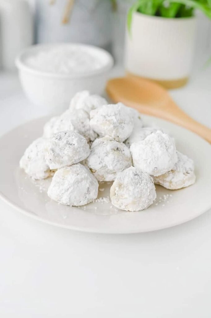 Freshly baked chocolate chip snowball cookies coated in powdered sugar stacked on a white plate.