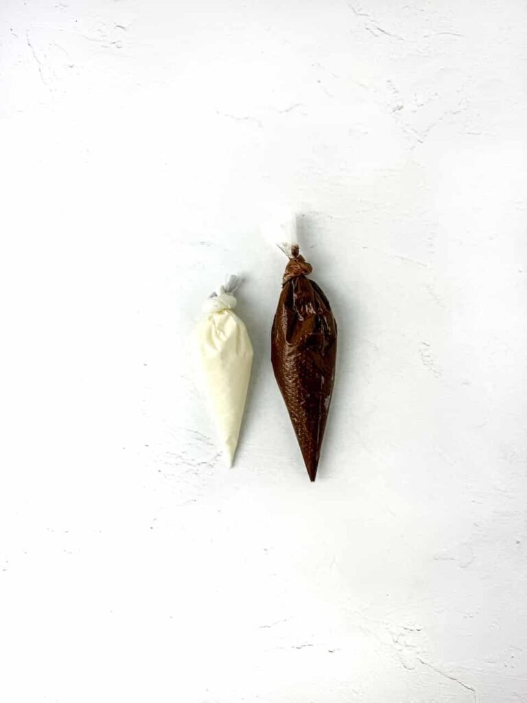 Two piping bags filled with melted white and brown chocolate resting on a white surface, prepared for decorating by piping the eyes, pupils, and beaks.